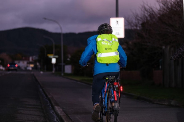 A woman rides a bike down a footpath in the dark; she's wearing a reflective backpack cover lit up by headlights