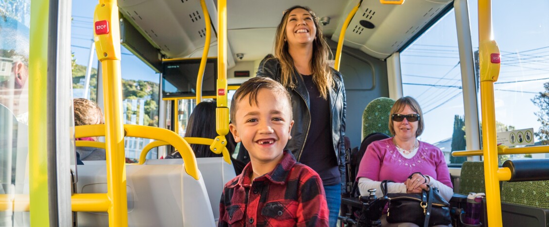 Inside a bus, a child and their mum smile as they walk past a woman in an electric wheelchair
