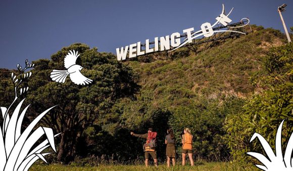 Three people standing at the base of the Miramar hill with the Windy Wellington sign, with flax and a pīwakawaka/fantail hand drawn at the corners