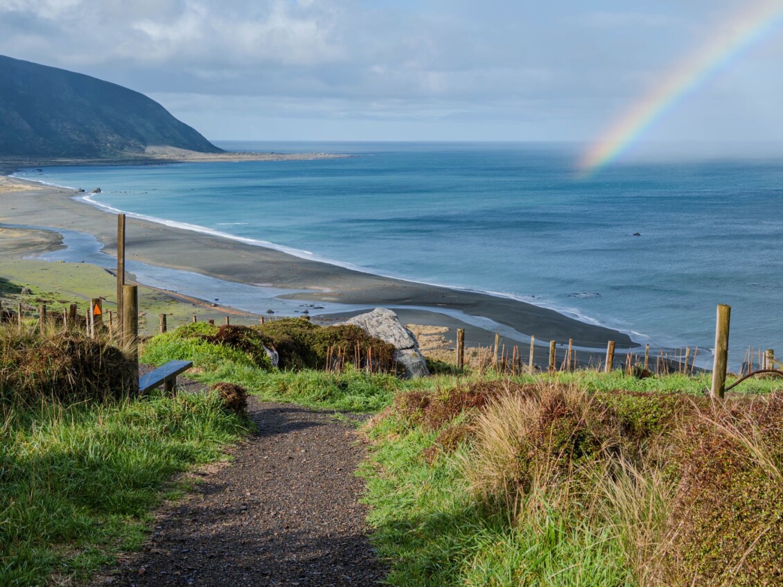 The view from the lightkeeper's cottages down to the beach at Baring Head on a cloudy but bright day, with a rainbow arcing over the sea