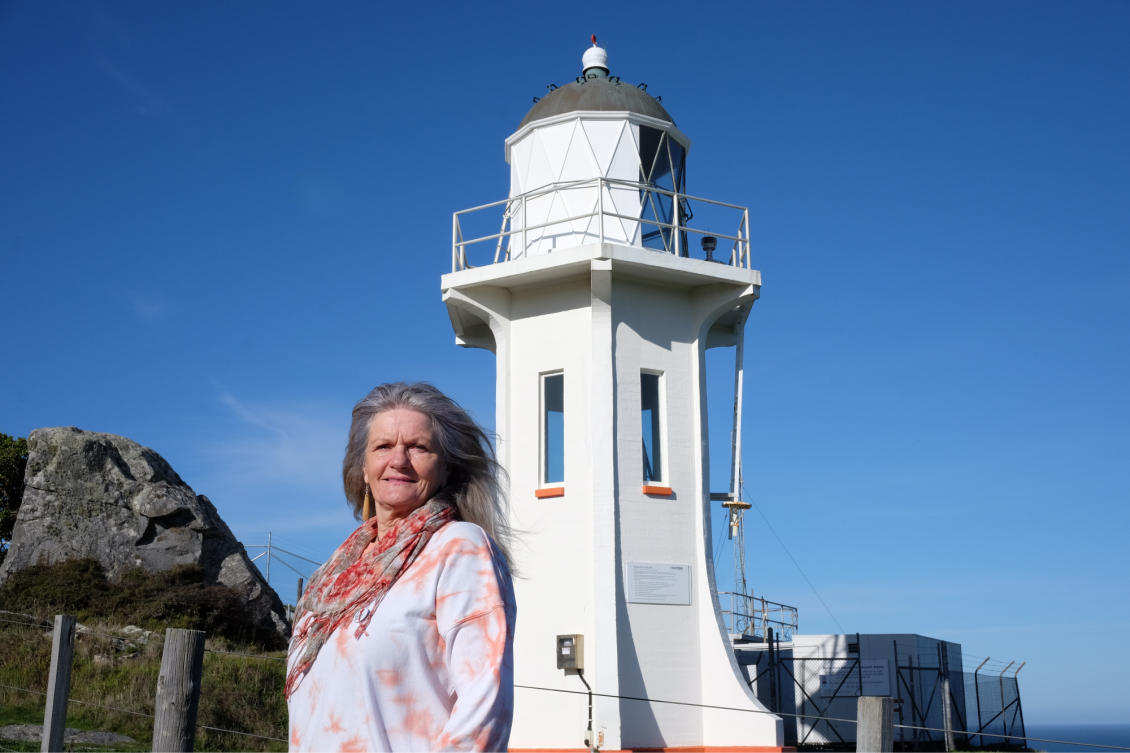 Gwen McDonald standing in front of the lighthouse at Baring Hea/Ōruapouanui on a bright sunny day
