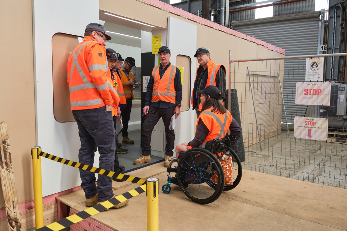 A group of people in hi-vis, five standing and one in a wheelchair, talk at the door to the Tūhono mock-up train