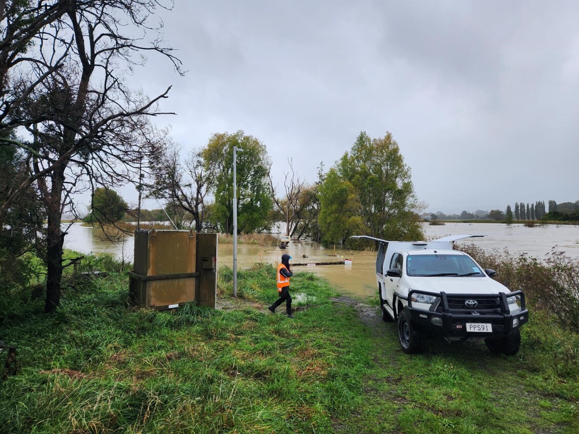 A flooded river surrounds the Waihenga Bridge site, with a Greater Wellington ute parked on grassy ground and a worker in high visibility clothing walking near rising water.